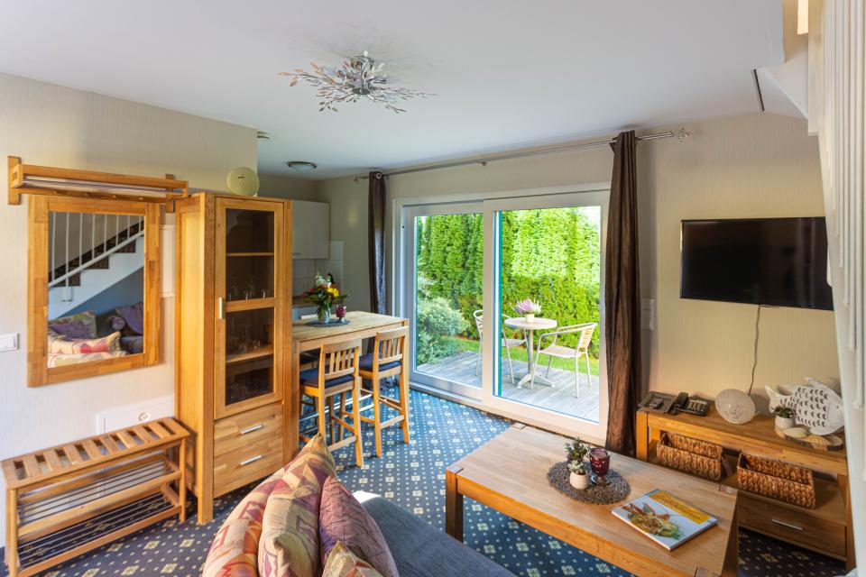 Apartment im Haus 2 Wohnzimmer mit Couchtisch, niedriges Sideboard, Fernseher an der Wand und Blick auf die Terrasse mit Tisch und 2 Stühlen
- nach rechts Blick in die kleine Kitchenette und erhöhtem Esstisch mit 2 Hochstühlen