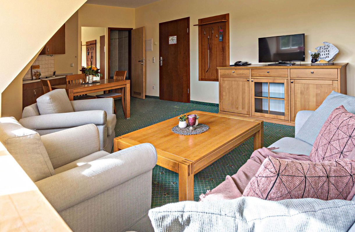 Apartment Fischhaus Wohnzimmer mit 2 sitzer Couch, Couchtisch,  2 Sesseln, 3 türige Kommode mit Fernseher darauf - links kleine Kitchenette mit Esstisch und 4 Stühlen